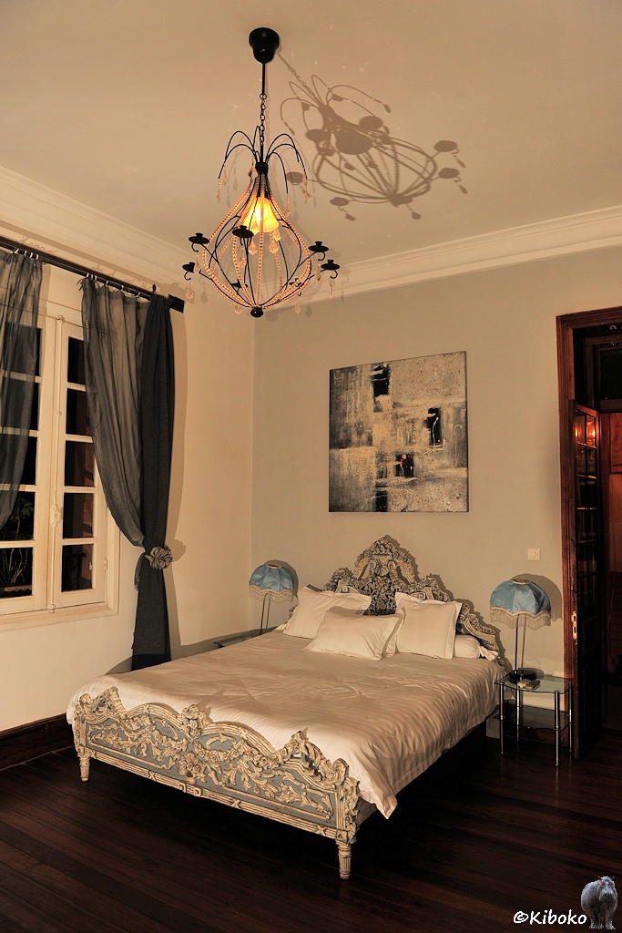 Das Bild zeigt eine Hochformataufnahme eines Hotelzimmers. Ein breites Bett mit silbernen, geschnitzen Giebeln steht unter einem Filigranen Kronleuchter, der an der Decke einen Schatten wirft. Die dunklen Vorhänge sind offen.