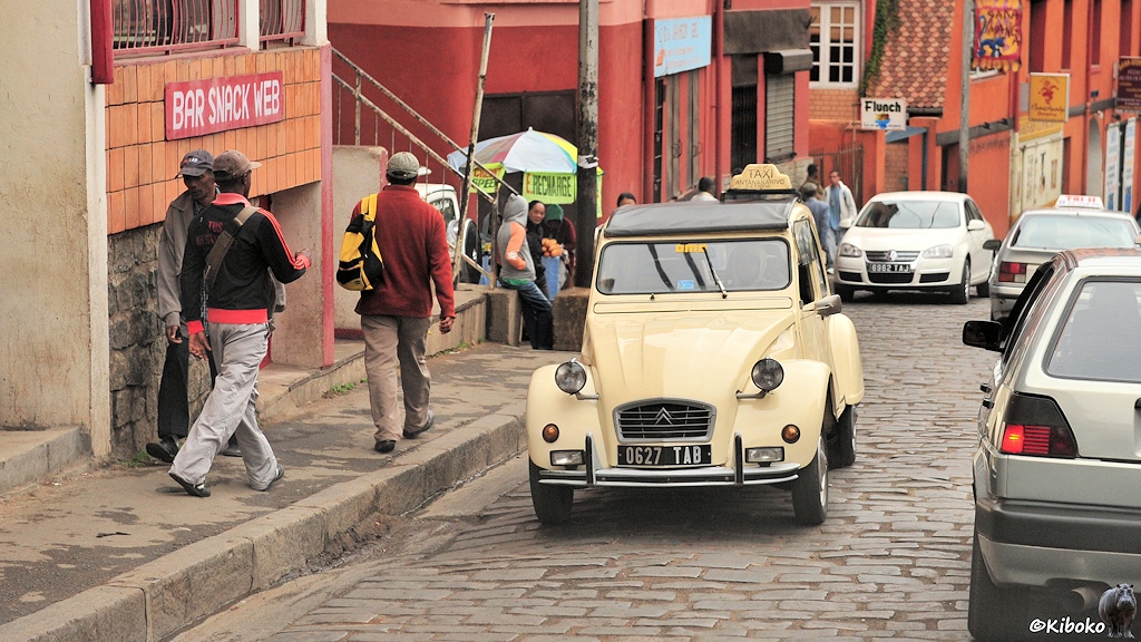 Das Bild zeigt ein entgegenkommendes beigefarbenes Taxi auf einer Pflasterstraße. Am linken Straßenrand stehen Häuser. Das erste Haus hat ein Schild mit der Aufschrift BAR SNACK WEB. Drei Männer laufen auf dem Gehsteig.
