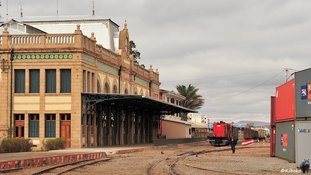 Das Bild zeigt eine rote Diesellokomotive vor einem Bahnhofsgebäude. Der einzige Bahnsteig ist am Gebäude mit einem Dach geschützt. Am rechten Bildrand steht eine Reihe Container.