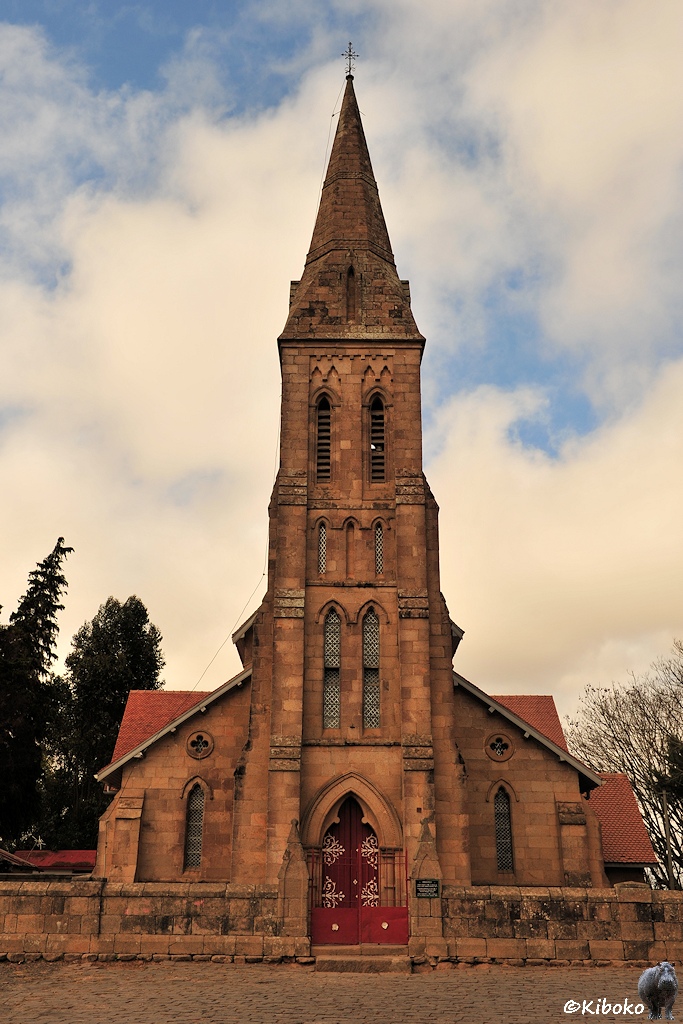 Das Bild zeigt eine Hochformataufnahme einer Kirche aus braunem Sandstein. Die Turmsptize ist sechseckig und ebenfalls aus braunem Sandstein. Die Kirche ist ungefähr dreimal so breit, wie der Turm.