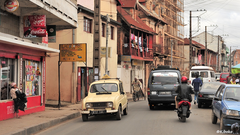 Das Bild zeigt ein entgegenkommendes beigefarbenes Taxi auf einer Städtischen straße. Link stehen Häuser. Am linken Bildrand lehnt jemand an einem Ladenfenster. Das Haus ist rot amgemalt mit den Buchstaben THB. Voraus fahren ein Moped und ein dunkelgrauer Kleinbus.