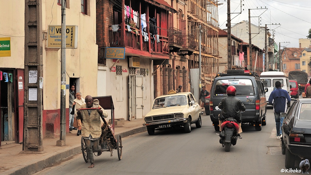 Das Bild zeigt eine beigefarbenes Taxi als Stufenhecklimousine in einer Straße mit dreistöckigen Gebäuden. Vor dem Taxi läuft ein Rikschafahrer. Voraus fahren ein Moped und ein grauer Kleinbus.