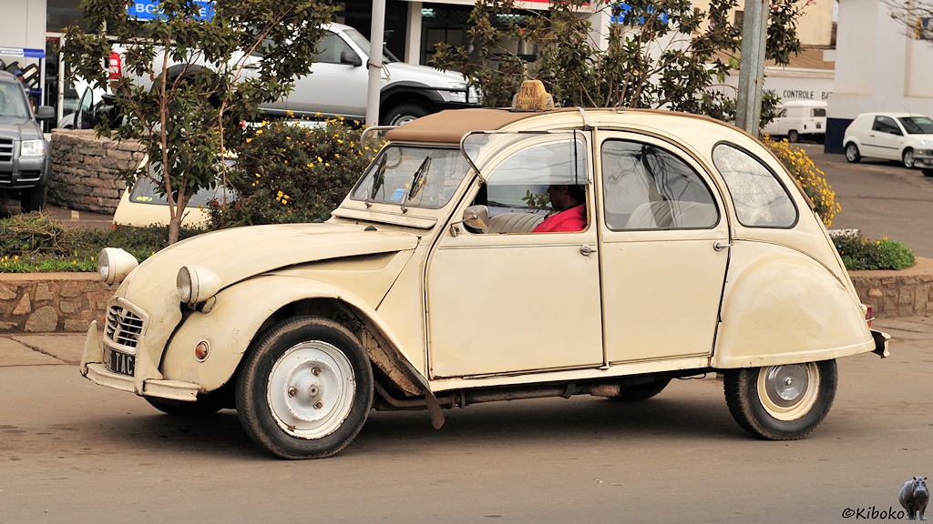 Das Bild zeigt einen beigefarbenen, alten franzöischen Kleinwagen Ente als Taxi von der Seite. Das Seitenfenster ist hochgeklappt. Der Fahrer im roten Hemd wartet auf neue Aufträge.
