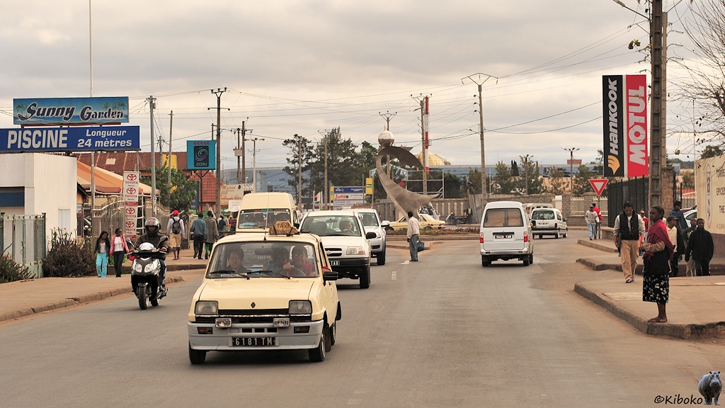 Das Bild zeigt ein entgegenkommendes kleines, beigefarbenes Taxi mit grauer Stoßstange auf einer breiten Teerstraße. An der Straße sind kleinere Läden und Werstätten. Im Hintergrund ist ein Kreisverkehr.