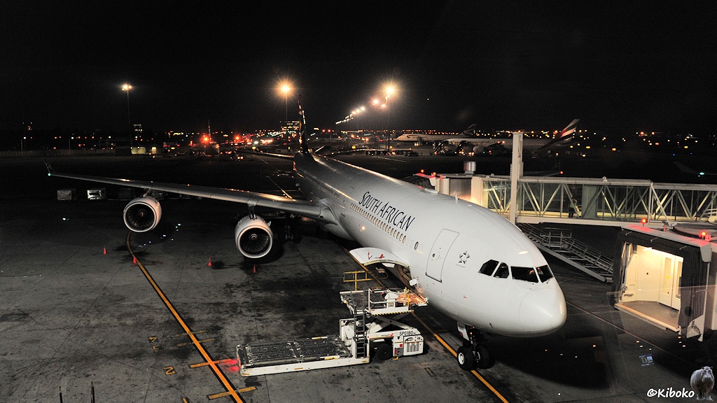 Das Bild zeigt ein weißes Düsenflugzeug mit vier Triebwerken nachts auf einem Flughafen. Die Gangway ist angedockt. Im Vordergrund wird mit einem Hublader Gepäck durch die offene Gepäcktür verladen.