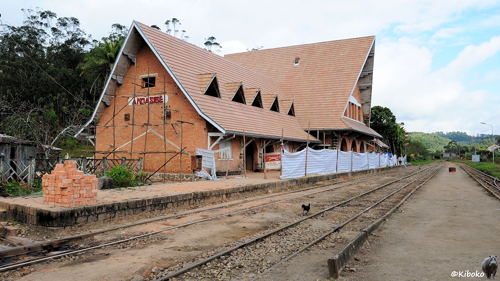 Ein Bahnhofsgbäude aus roten Ziegelsteinen mit markantem Spitzdach steht an einem dreigleisigen Bahnhof und wird gerade renoviert. Eine Zaun aus weißer Plane auf den Hausbahnsteig grenzt die Baustelle ab.