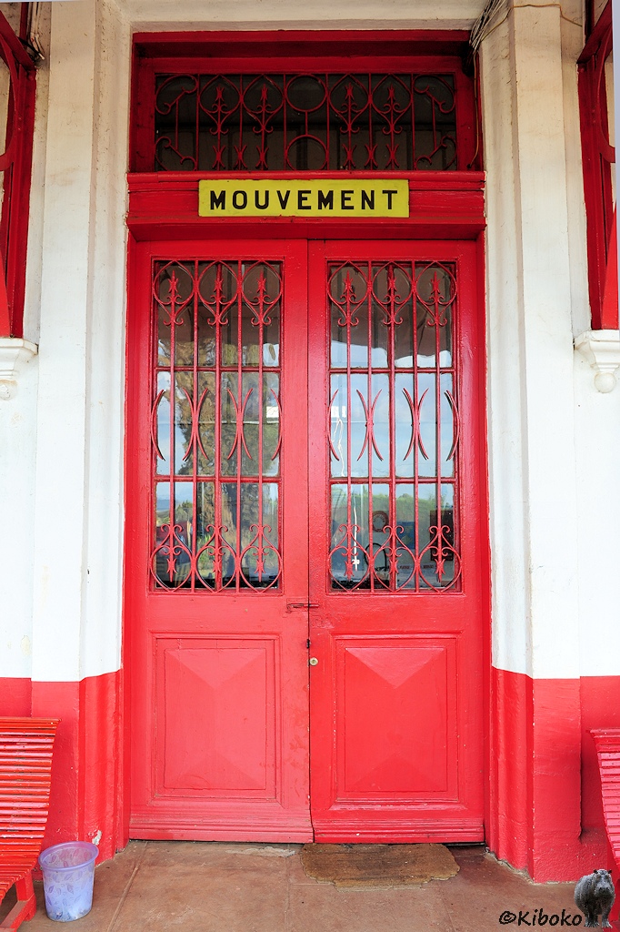 Das Bild zeigt eine hohe rote Tür im Hochformat mit vergitterten Glasfenster in der oberen Hälfte. Über der Tür ist ein gelbes Schild mit schwarzer Schrift auf dem steht Mouvement.