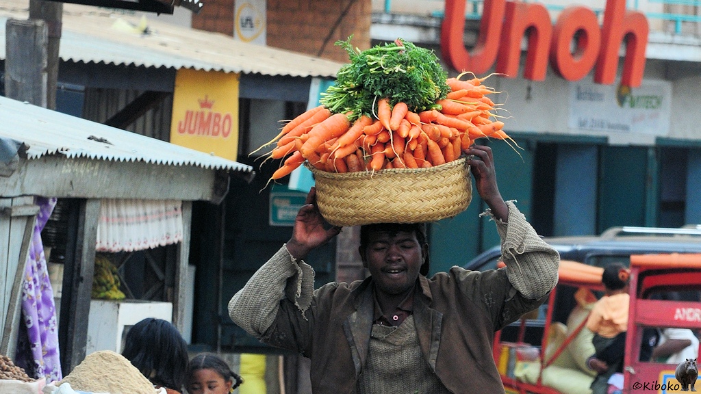 Das Bild zeigt einen Mann der auf dem Kopf einen Strohkorb mit Karotten trägt .