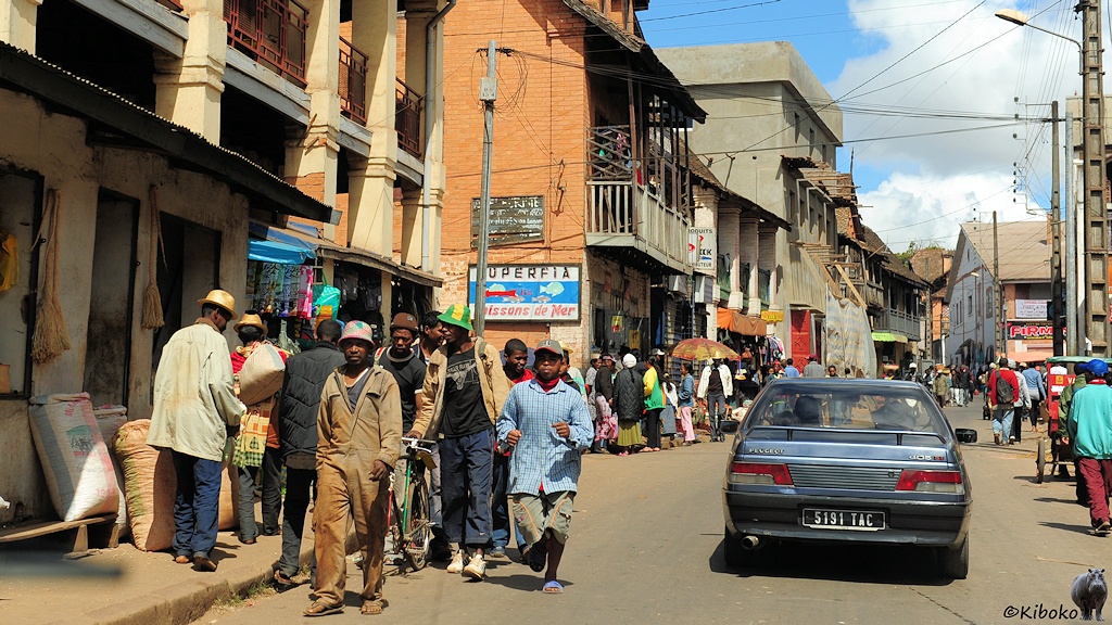 Das Bild zeigt eine Geschäftsstraße mit Läden in zwei- und dreigeschossigen Häusern. Viele Menschen laufen auf der Straße. Voraus fährt ein PKW in blau metallic.