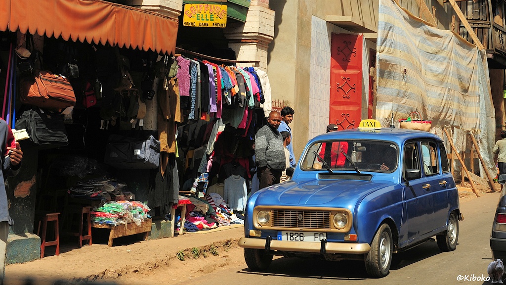 Das Bild zeigt einen entgegenkommenden Kleinwagen vom Typ R4 in blaumetallic mit Taxi Schild. Links ist ein Klamottenladen mit bunter Wäsche an Stangen und auf Wühltischen.