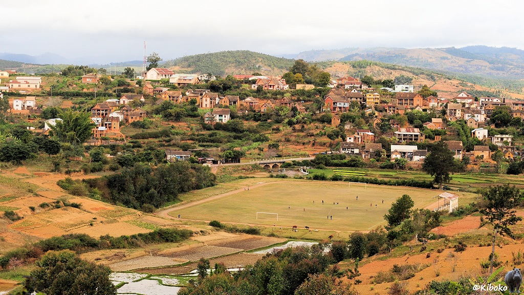 Das Bild zeigt weitläufige Aufnahme eines Ortes an einem Hang. Im Tal ist ein Fußballplatz.
