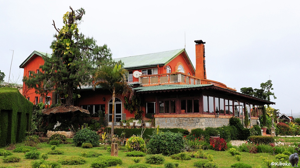 Das Bild zeigt ein zweistöckiges orangerotes Gebäude mit grünem Blechdach in einem gepflegten Garten. Am Haus ist eine große verglaste Veranda.