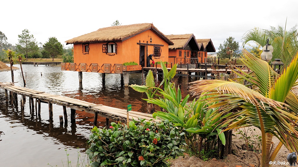 Das Bild zeigt drei kleine orange gestrichene Häuschen mit Schilfdach. Die Häsuchen stehen auf Stelzen auf einem See.