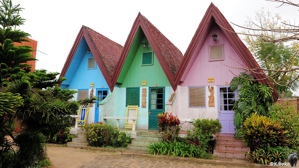 Das Bild zeigt drei aneinandergereihte Holzhäuschen mit sehr hohen spitzen Dächern. Die Häuse sind blau, grün udn violett angemalt.