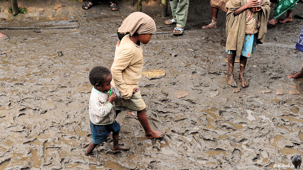 Das Bild zeigt zwei Kinder. Ein Mädchen mit einen kleinen Jungen, wie sie barfuß durch den Schlamm laufen.