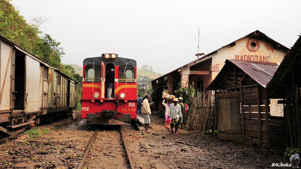 Das Bild zeigt die Zuglok direkt von vorn. Links sehen zwei gellgraue, gedeckte Güterwagen. Rechts ist hinter zwei Bretterbuden das Bahnhofsgebäude zu sehen. Männer verlassen mit Sack und Pack den Bahnsteig.