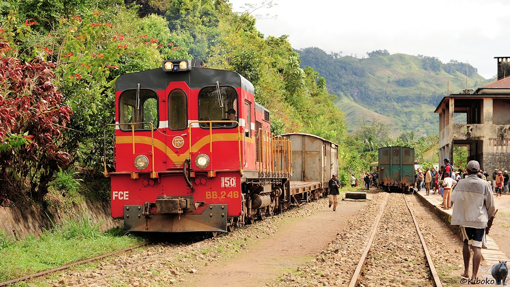 Das Bild zeigt eine rote Lokomotive mit gelben Zierstreifen. Sie hat einen flachen und einen gedeckten Güterwagen auf dem Nebengleis gegenüber dem Bahnhof abgestellt. Auf dem Hauptgleis steht der abgestellt Zug mit dem grünen Gepäckwagen.