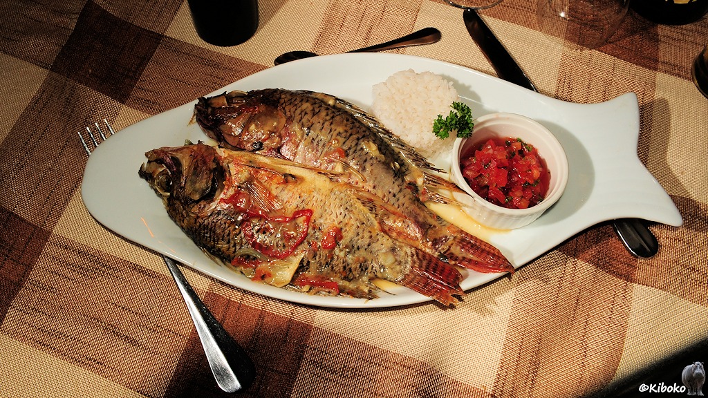 Das Bild zeigt zwei gebratene Fische auf einem Teller in Fischform. Dazu gibt es Reis und Paprika.