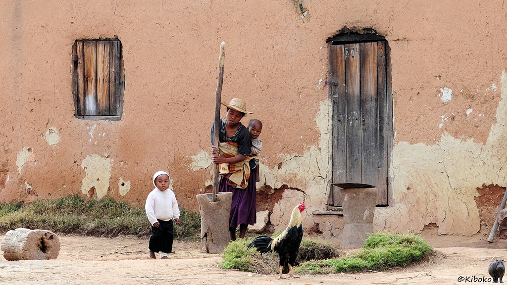 Das Bild zeigt eine Frau mit Strohhut vor einem rotbraunen Haus. Sie hat ein Baby auf dem Rücken. ein zweites Kind schaut zu, während sie mit einem langen Knüppel in einem ausgehöhlten Baumstamm stampft. Ein weiß-schwarzer Hahn steht daneben.