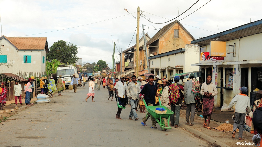Das Bild zeigt eine dichtbevölkerte Straße. Ein Mann schiebt eine grüne Schubkarre. Rechts sind kleine Läden. Waren und Säcke liegen auf beiden Straßenseiten auf dem Boden.