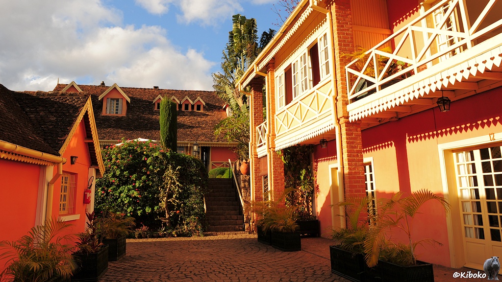 Das Bild zeigt zweistöckige Gebäude mit rotbraunen Wänden, Ziegelpfeilern und weiß gestrichenen Geländern und Fensterrahmen. Eine Treppe verbindet Gebäude auf mehreren Ebenen. Der Hof ist gepflastert.