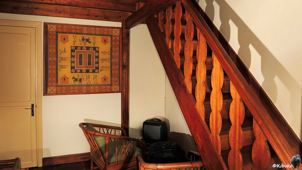 Das Bild zeigt einen Ausschnitt aus dem Hotelzimmer mit zwei Holzsesseln und einem kleinen tisch auf dem ein schwarzer Röhrenverseher steht. Eine Holztreppe fürht am rechten Bildrand nach oben.