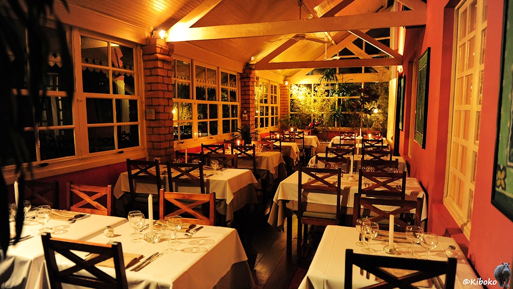 Das Bild zeigt einen Restaurantanbau mit 9 Tischen und abendlicher Beleuchtung. Die Tische sind gedeckt. Teilweise brennen weiße Kerzen.