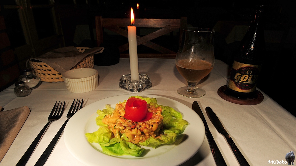 Das Bild zeigt die Vorspeise mit Nudelsalat, darum ausgelegten grünen Salatblättern und einer zur Rose geschnittenen Tomate. Im Hintergrund steht ein halbvolles Bierglas und eine Bierflasche mit der Aufschrift: Gold. Eine Kerze brennt auf dem Tisch.