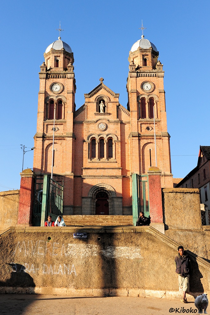 Das Bild zeigt das Portal einer aus Ziegelstein gemauerten Kirche mit zwei Türmen hinter einem geöffneten grünen Gittertor.Davor sitzen Kinder und ein junger Mann lehnt an einer Mauer.