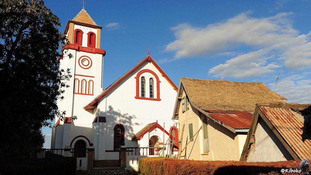 Das Bild zeigt weiße Kirche mit seitlichem viereckigen Turm. Die Fensterrahmen und Verzierrungen sind rot lackiert. Daneben stehen zwei kleinere hellbraune Häuser mit Spitzdach.