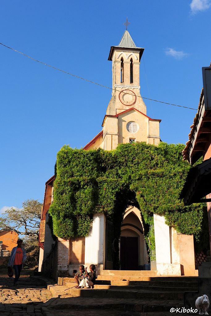 Das Bild zeigt eine hellbraune Kirche mit einem hohen viereckigen Turm. Der Weg zur Kirche führt durch ein mit Rankpflanzen überwucherten Tor mit Spitzbogen.