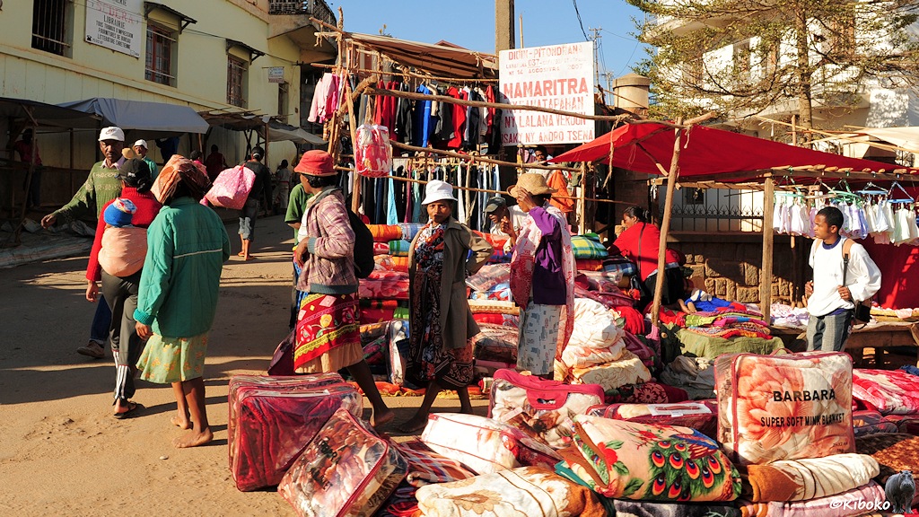 Das Bild zeigt einen Haufen bunter Decken, die in durchsichtigen Platiktüten verpacket sind. Dhinter ist eine Gruppe Frauen und weitere Marktstände mit Decken und Klamotten.