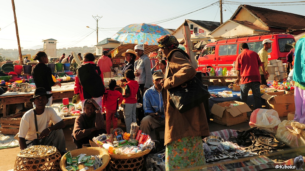 Das Bild zeigt einen Marktplatz mit vielen kleinen Ständen. Im Vordergrund sind drei Männer die aus drei Körben ihre Waren verkaufen.