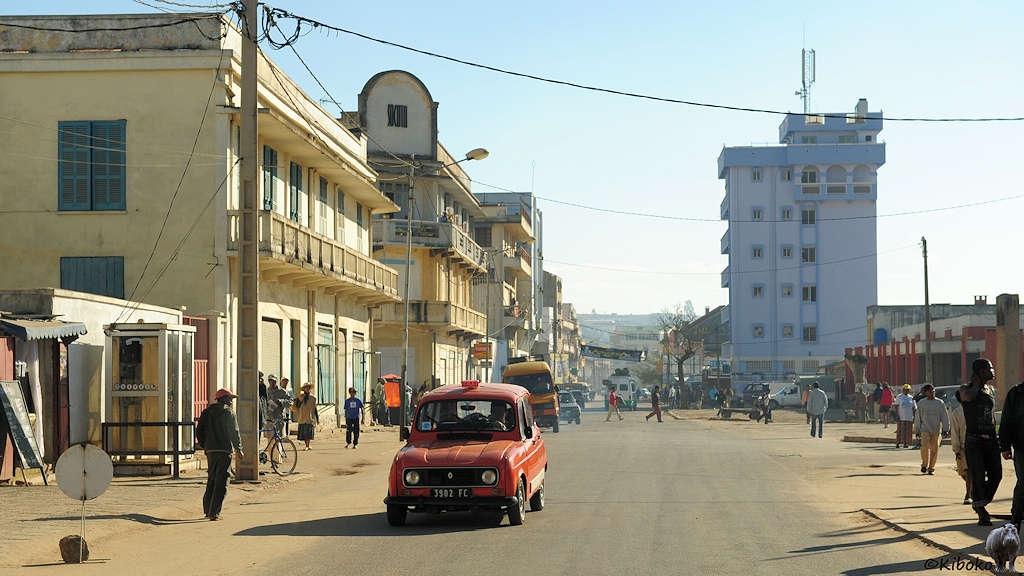 Das Bild zeigt eine breite, staubige Teerstraße. Ein orangeroter R4 kommt als Taxi entgegen. An der Straße stehen mehrgeschossige Häuser.