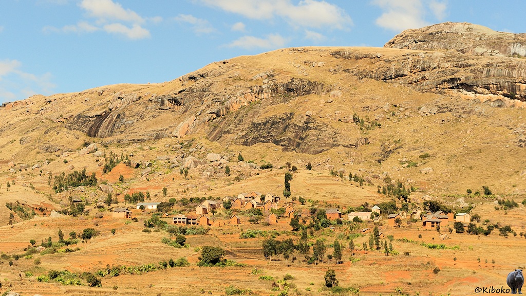 Das Bild zeigt ein Dorf aus kleinen erdfarbenen Häusern und mit einer Kirche an einen Berghang. Hinter dem Dorf wird der Berghang zu steilen Felsen. Bäume stehen nur vereinzelt.