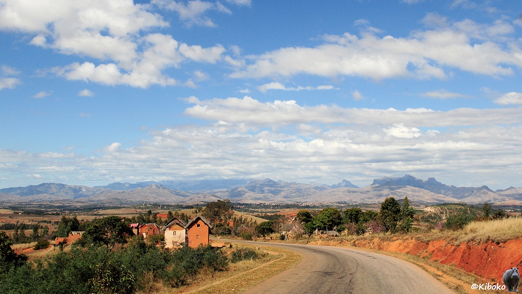 Das Bild zeigt eine weite Landschaftsaufnahme. Die Straße führt in einen großen Linksbogen durch eine Ortschaft. Dahinter ist ein weites Tal mit einer einer Ortschaft vor einer Bergkette.