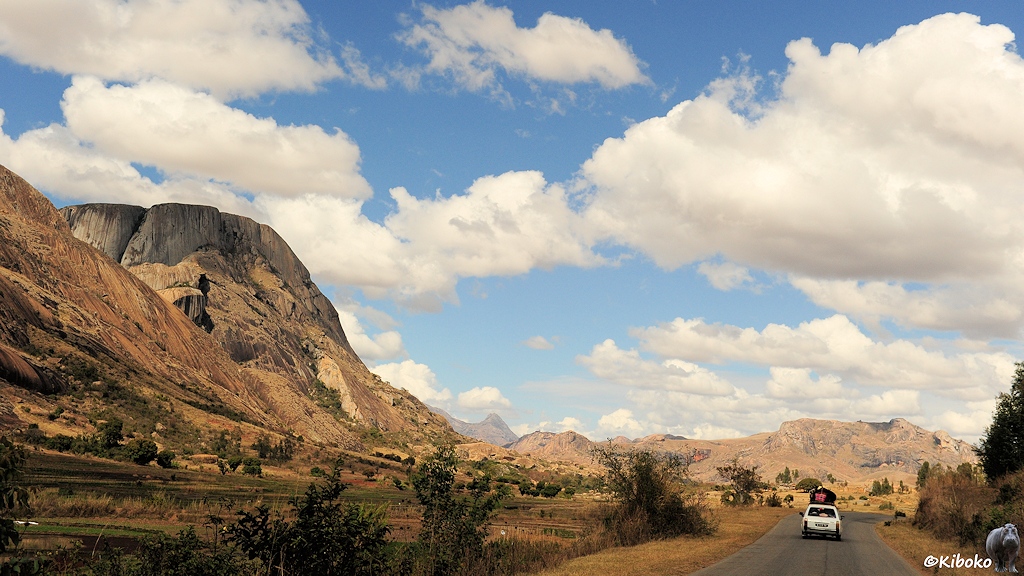 Das Bild zeigt ein Tal in einer Gebirgslandschaft mit hohen Bergen aus rundgeschliffenen grauen Fels. Auf der Straße fährt vor uns ein überladener weißer Kombi. Einzelne Wolken ziehen über den Himmel.