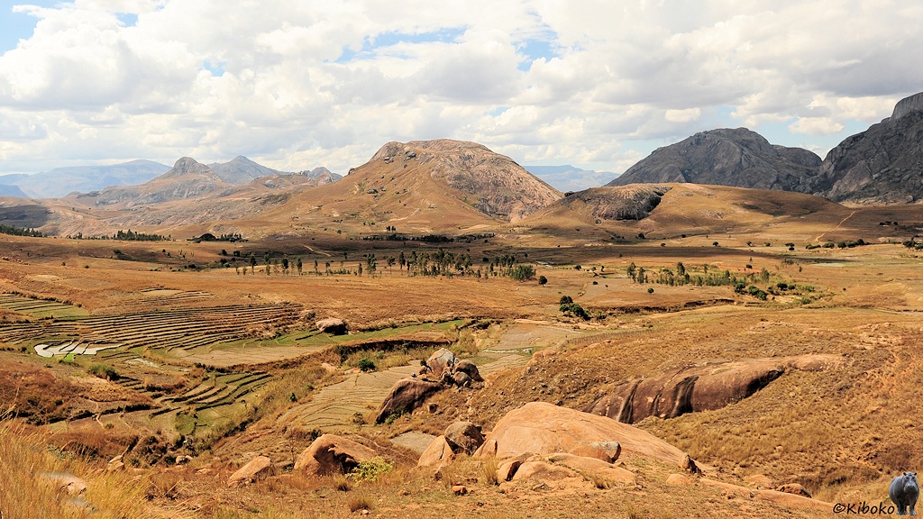 Das Bild zeigt einen Blick von einer Anhöhe in ein weites Tal. Im Vordergrund leigen Felsen. Im Tal sind Felder. Der Boden ist graubraun. Im Hintergrund sind mehrere Bergketten.