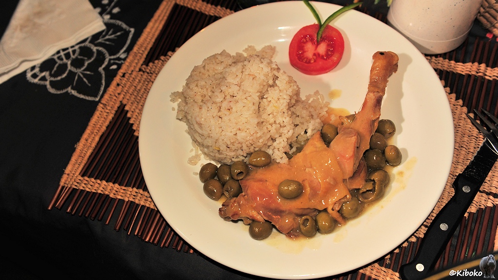Das Bild zeigt eine Hähnchenkeule mit grünen Oliven, einen Reisklumpen und eine Dekorationstomate.