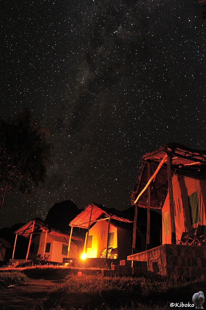 Das Bild zeigt drei Hauszelte im Hochformat. Vor dem mittleren Zelt steht eine Petroliumlampe. Über den zelten geht senkrecht die Milchstraße in den Himmel.