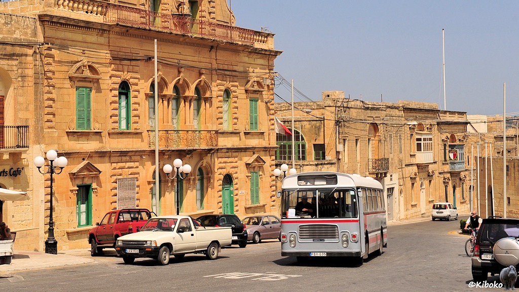 Das Bild zeigt einen grauen Bus mit weißem Dach und roten Zierstreifen in einer Straße mit Gebäuden in hellbraunen Sandstein.