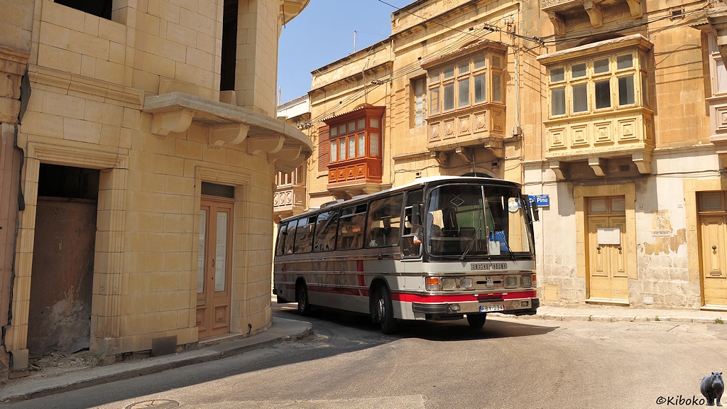 Das Bild zeigt einen grauen Bus mit großen Frontscheiben fährt durch eine enge Kurve zwischen hellbraunen Häusern mit gemauerten Balkonen.