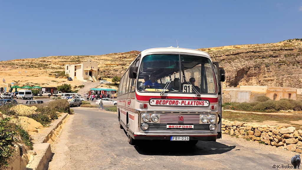 Das Bild zeigt einen weißen Bus mit roten Zierstreifen und vor einem beigen Felskulisse mit einer kleinen Kirche.