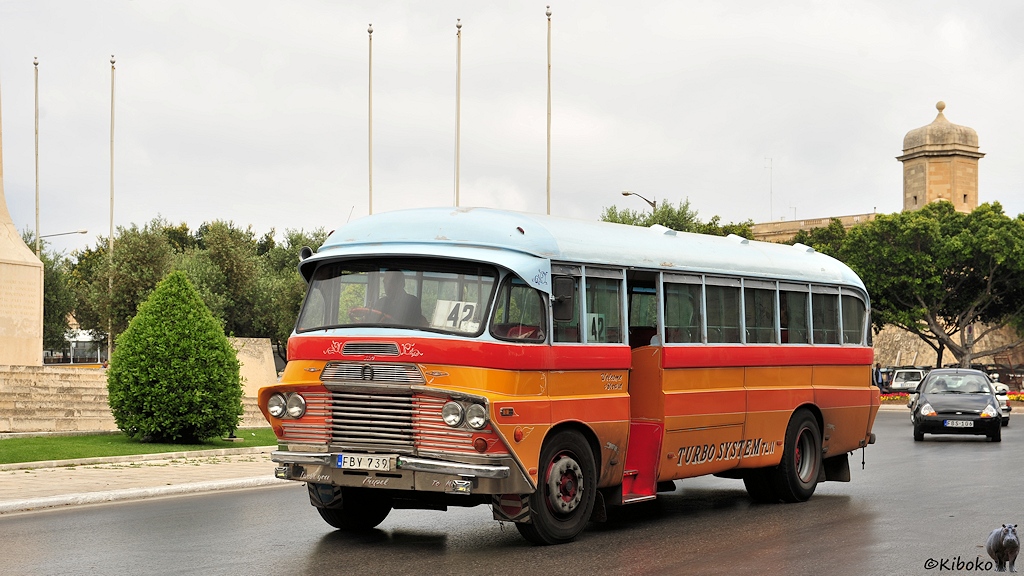 Das Bild zeigt einen alten gelben Bus mit hellblauem Dach, roten und orangenen Zierstreifen, waagerechte vercrohmte Zierleisten an der Front und der Aufschrift Turbo System an der Seite. Im Hintergrund ist ein Turm aus Sandstein.