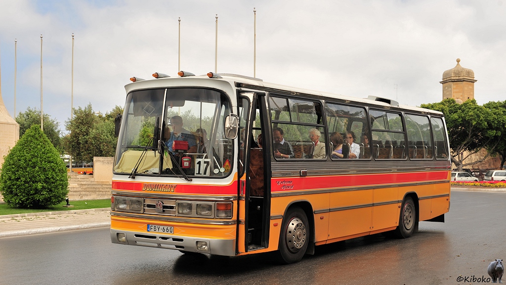 Das Bild zeigt einen besetzten moderneren gelben Bus mit sehr großen Frontscheiben. Auch hier ist der Sandsteinturm im Hintergrund.