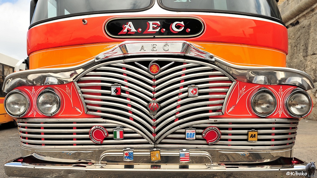 Das Bild zeigt einen verchromten Kühlergrill mit zahlreichen verchromten Schildern von Automobilclubs und Hufeisen. Darüber ist der Schriftzug A.E.C.