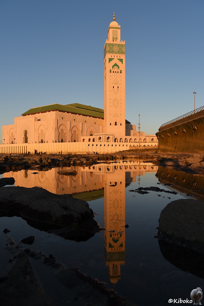 Das Bild zeigt eine Moschee mit viereckigen Turm im Abendlicht im Hochformat. Der Turm spiegelt sich in einen Wasserfläche zwischen Felsen am Meer .
