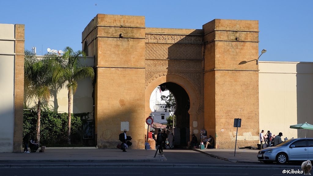 Das Bild zeigt ein Stadttor aus Sandstein mit seitlichen Türmen. Das Tor führt durch eine beigefarbene hohe Stadtmauer.