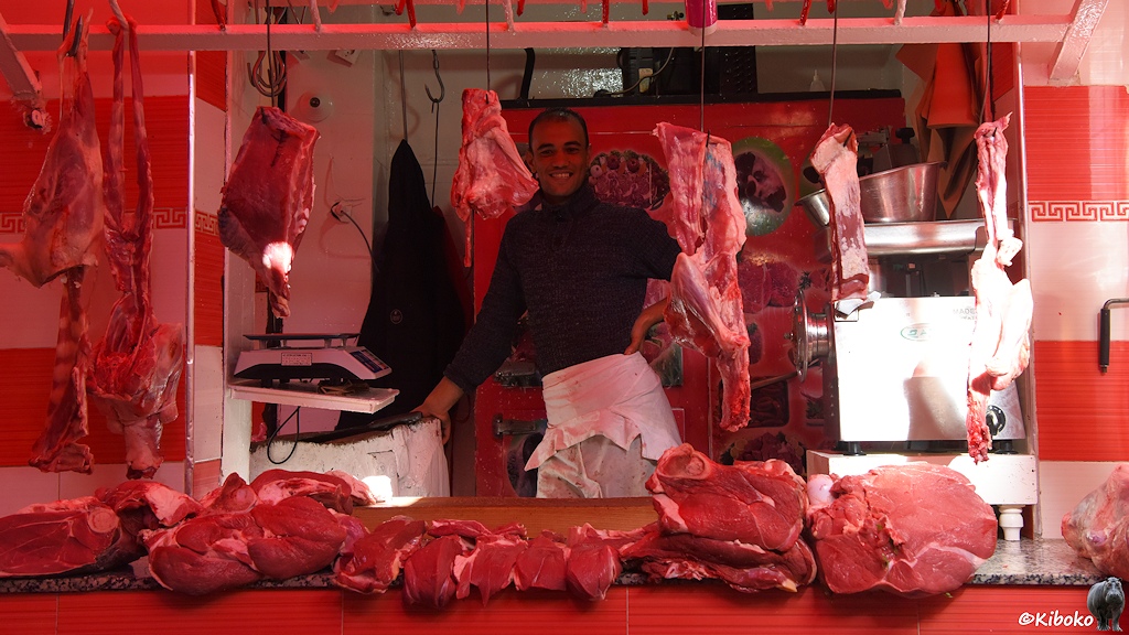 Das Bild zeigt einen jungen, lächelnden Fleischverkäufer. Große rote Fleischbrocken liegen in der Auslage im Schaufenter. Weitere Fleischbrocken hängen an Haken von der Decke. Un Geschäft steht eine Verkaufswaage und ein Fleischwolf.