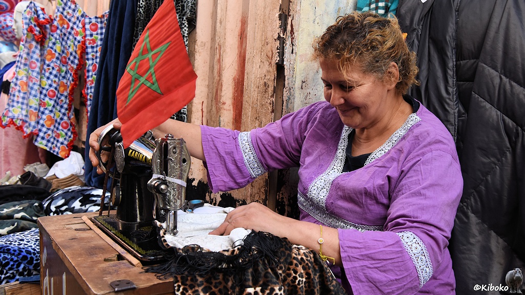 Das Bild zeigt eine ältere Frau in einer lilafarbenen Bluse vor einer schwarzen Nähmaschine mit der Aufschrift Karina. Über der Nähmaschine hängt eine rote Fahne mit grünem Stern.